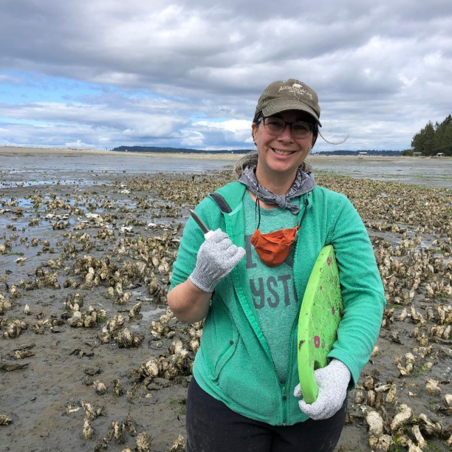 Melissa Poe holds an oyster shucking knife and cutting board, ready to harvest and shuck shellfish on a mudflat.