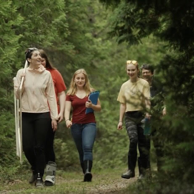 A group of students walk through a wooded area. Students carry a clipboard and other scientific data collection tools.