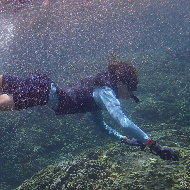 Samuel swims in snorkeling gear near to the water's surface against a background of rock and corals. The leg of a second snorkeler can be seen as the person swims out of the shot. 