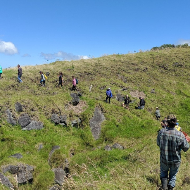Students and educators hike along a grassy hillside in a single-file line.