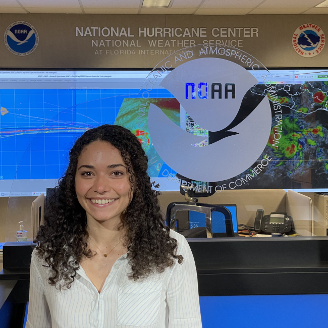 Delián poses in business casual clothing and smiles for the camera in front of a glass wall with an etched NOAA logo on it. In the background, the words "National Hurricane Center, National Weather Service" are written on the wall above two large monitors showing satellite weather
