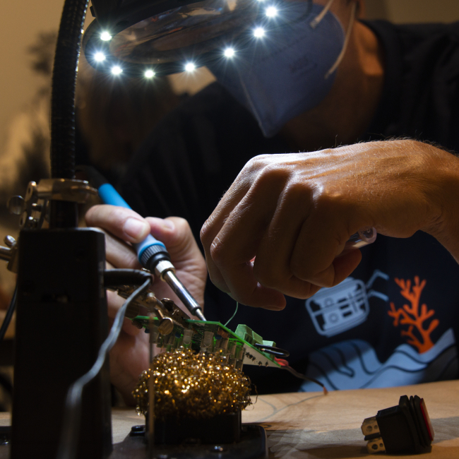  Teacher at Sea alumnus Jeff Miller sits at a workbench, his face mostly obstructed from view by a large magnifying glass ringed with bright lights. A small, square green circuit board is mounted at an angle underneath the light. The focus of the photo is on Jeff’s hands. With his right hand, he holds a soldering iron to the circuit board. His left hand steadies a thin wire against the board while grasping a tube of solder. Other project components — a switch, a brass wool scouring pad — are visible on the 