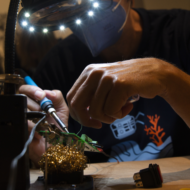 Teacher at Sea alumnus Jeff Miller sits at a workbench, his face mostly obstructed from view by a large magnifying glass ringed with bright lights. A small, square green circuit board is mounted at an angle underneath the light. The focus of the photo is on Jeff’s hands.