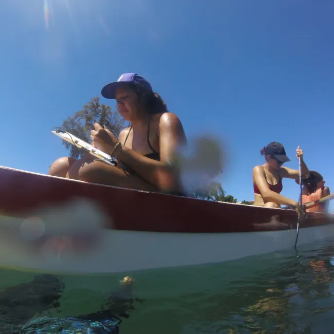 A woman wearing a hat and sitting on a canoe, taking notes while two other women sitting behind her row and advance the canoe on the water.