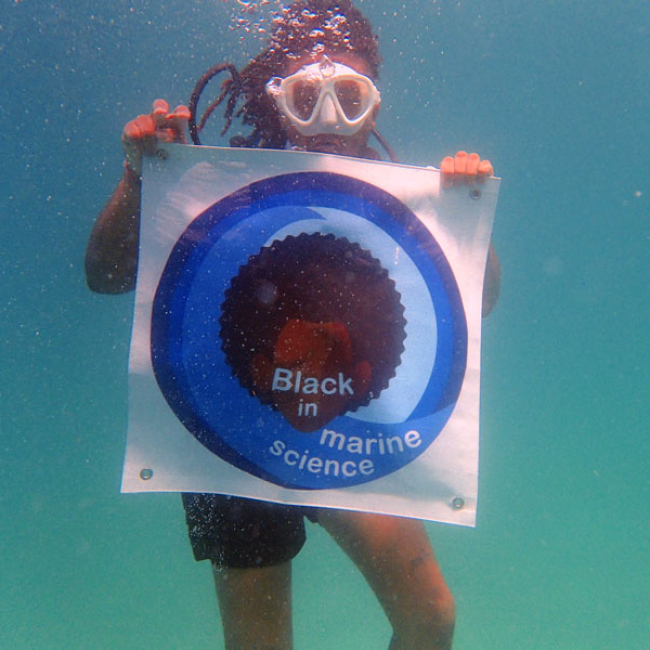 A young black student underwater scuba diving and holding up a sign with the Black in Marine Science logo.
