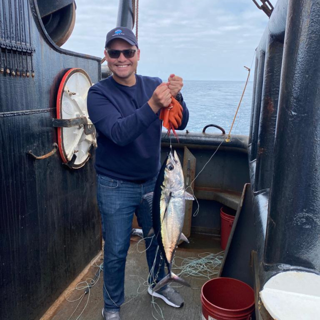 Michael Gutiérrez Santiago stands posing aboard NOAA Ship Bell N. Shimada while holding up a big Albacore Tuna by a string.