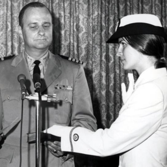 A black and white photo of Ensign Pamela Chelgren being sworn in by Secretary of Commerce Peter G. Peterson on July 6, 1972. Her father, Navy Captain John Chelgren, stands between them, holding the Bible for the ceremony.