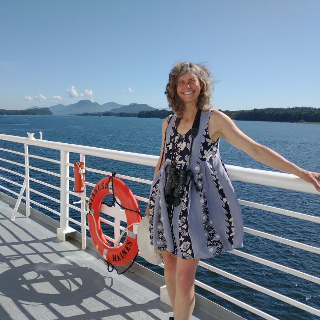 Denise wears a sundress and stands against the railings of the ferry and smiles. In the background is the beautiful coastline and mountains of Alaska.