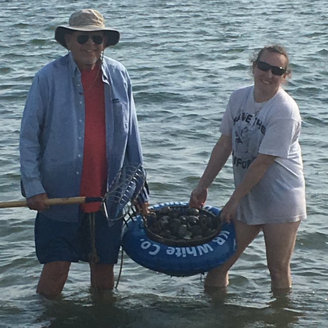 Joseph Abromaitis and Grace Simpkins stand knee-deep in water holding a basket of quahogs and smiling.