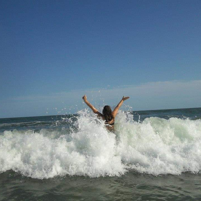 Kayla has her back to the camera and holds her arms in the air as she jumps into a large ocean wave.
