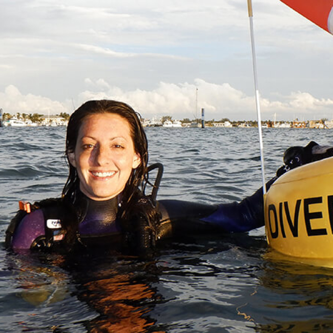 Rachel is at the surface of the water with her hand on a large flotation device that says “Diver below.” The dive flag is attached to the top of the inflatable device.