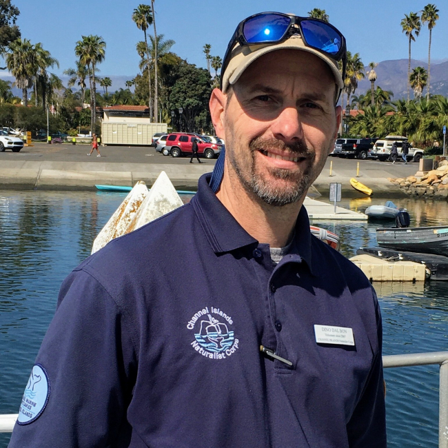 Dino stands on a boat, just a few yards from the pier, wearing a blue shirt that bears the logo of the Channel Island Naturalist Corp.