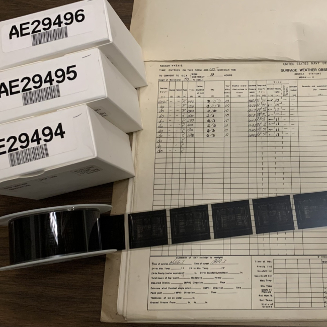 A reel of microfiche is unrolled on top of a Naval weather logbook alongside boxes of other reels.