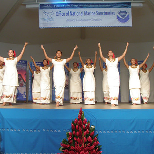 Samoan dancers lift their arms on stage below a National Marine Sanctuary sign.