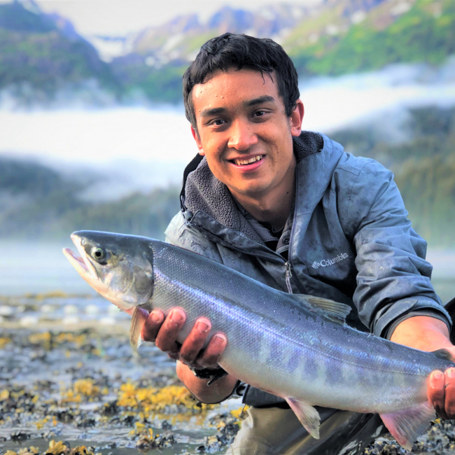 Andrew Tokuda, Global environmental science major at University of Hawaiʻi at Mānoa and 2018 NOAA Hollings scholar, holds a chum salmon while kneeling beside the Tutka Bay estuary. Tokuda interned at the Kachemak Bay National Estuarine Research Reserve in Homer, Alaska, in summer 2019.