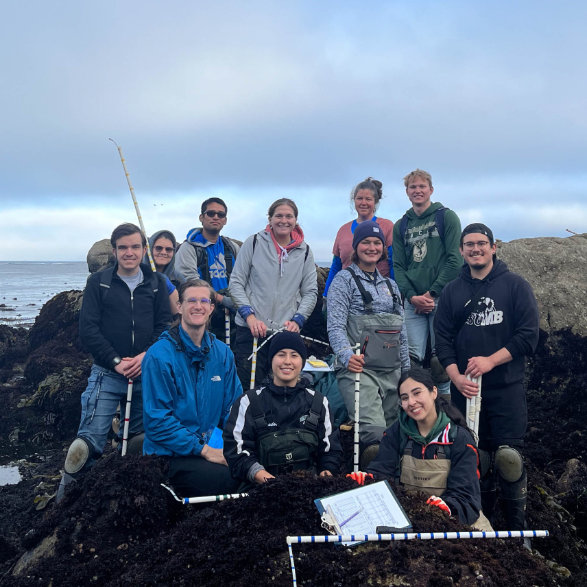 A group of people  in cold water field clothes hold sampling gear and pose in an intertidal area. It is rocky with pools of water and the lower portions of the rocks are covered in ocean vegetation as if they are frequently underwater. 